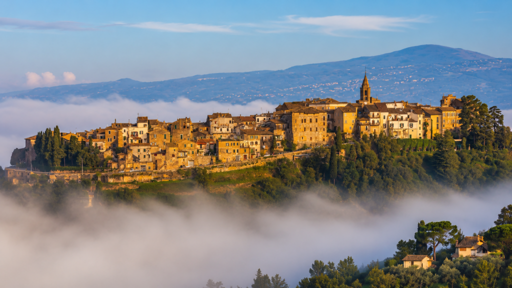 Veduta panoramica di Collevecchio, borgo della Sabina Tiberina, con case storiche, campanile, colline verdi e nebbia sulla Valle del Tevere.